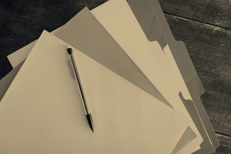 A fanned stack of labelled file folders on a wooden desk with a pen resting on top